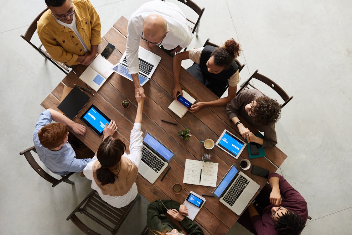 an overhead shot of several people sitting around a wooden table. They each have their laptops out. Two people are reaching across the table to shake hands.