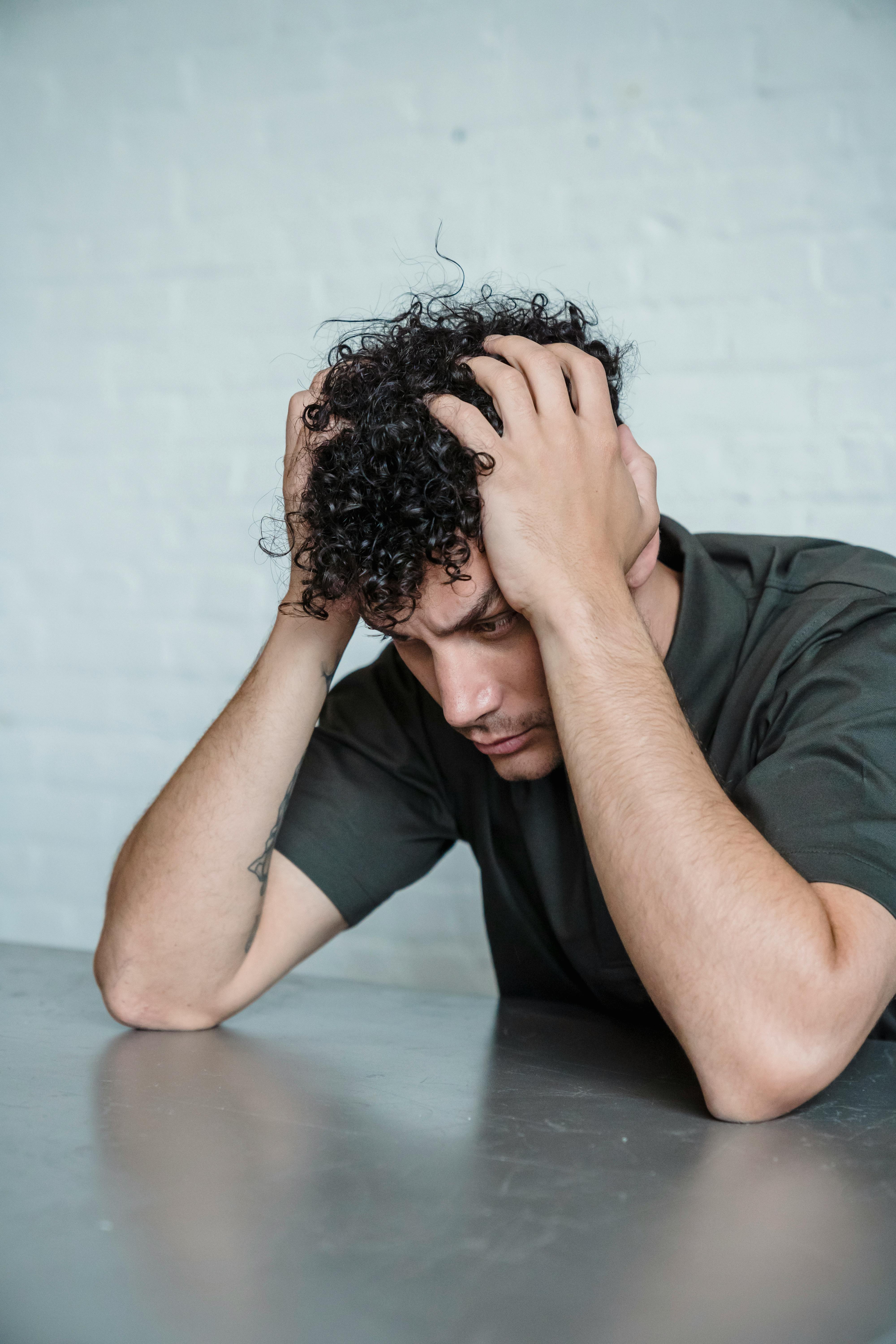 A man sitting at a metal table looking stressed with his hands on his head.