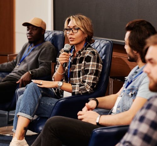 A woman with light hair and black framed glasses speaking from a chair. A man in the background sits in a similar chair.