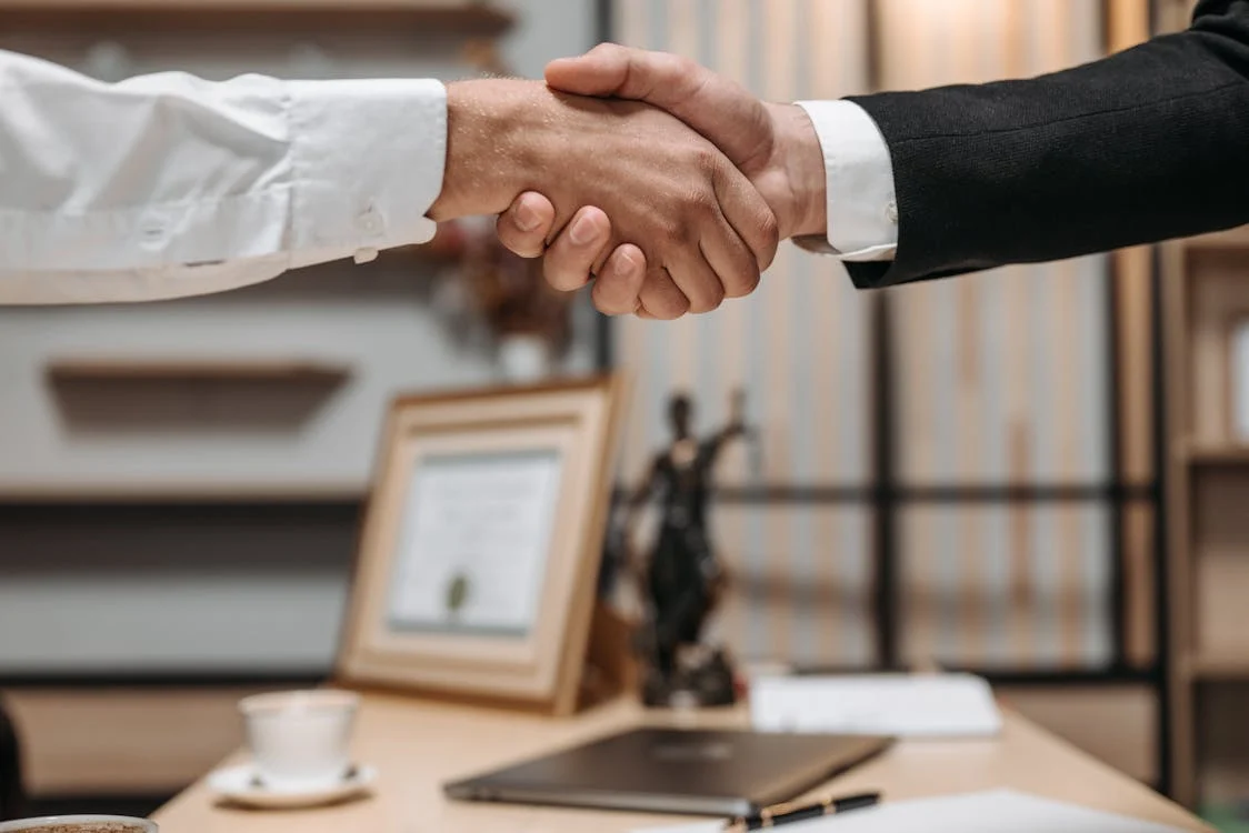 A picture of a handshake over a desk with a closed laptop and a cup of tea or coffee. One person is wearing a dress shirt and the other is wearing a suit jacket.