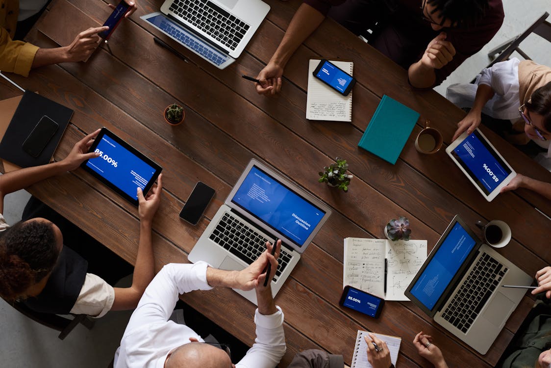 An overhead picture of a group of people sitting at a brown wooden table. Each one is using a computer or smart device with a blue screen with text displayed.