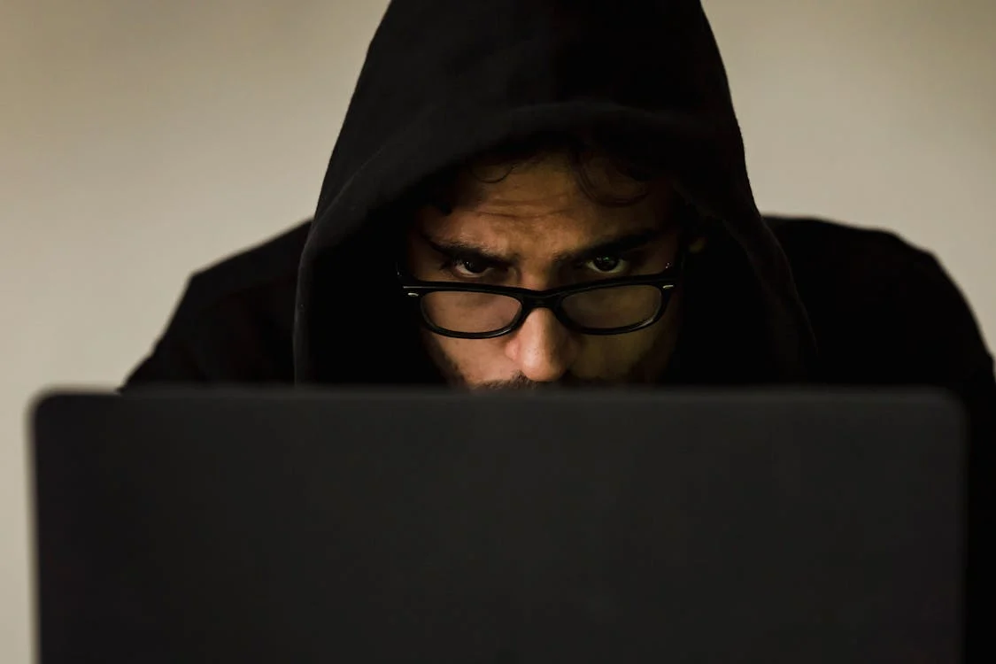 A man with an intense, focused look on his face sits behind his laptop computer with his hoody pulled up over his head.