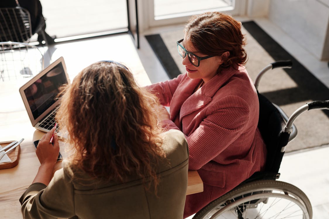 Two women working on a computer together. One is wearing pink, has glasses, and is sitting in a wheelchair. The other is wearing an olive green shirt and has her back to the camera.