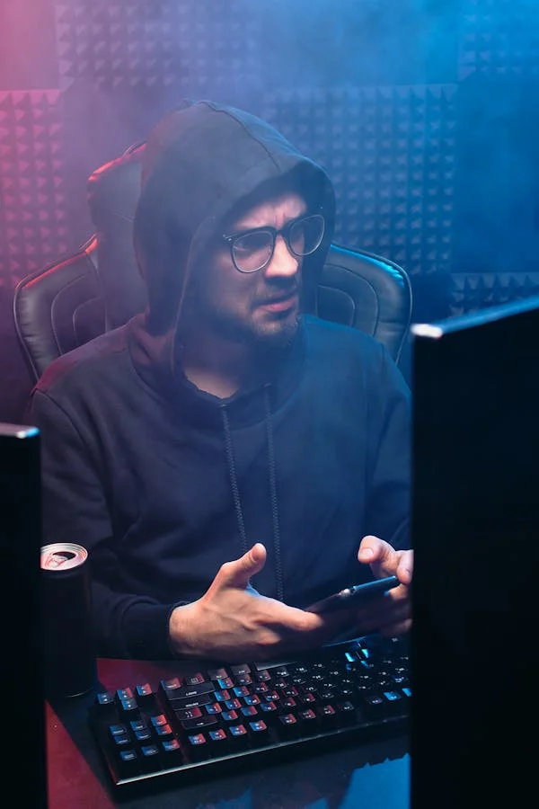 A bewildered and distressed looking man sits in a hazy room behind a pair of computer monitors. He has his phone in his hand.